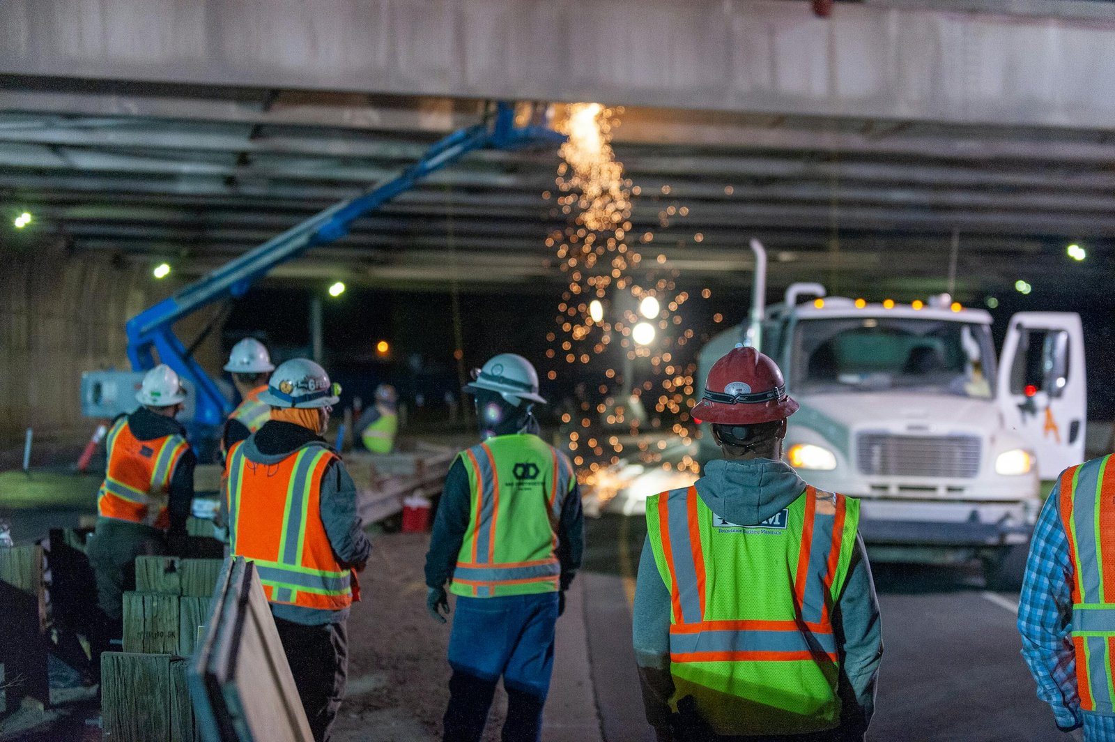 Nighttime road construction with workers under a bridge, sparks flying from machinery.